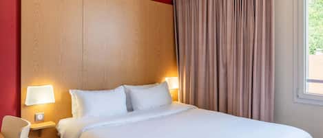 A hotel room with a large bed, two bedside lamps, a wooden headboard, a window with curtains, and a small table with a chair.