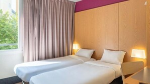 A hotel room with two beds, a wooden headboard, a window with curtains, and bedside lamps. - B&B HOTEL Lyon Caluire Cité Internationale (Caluire-et-Cuire)