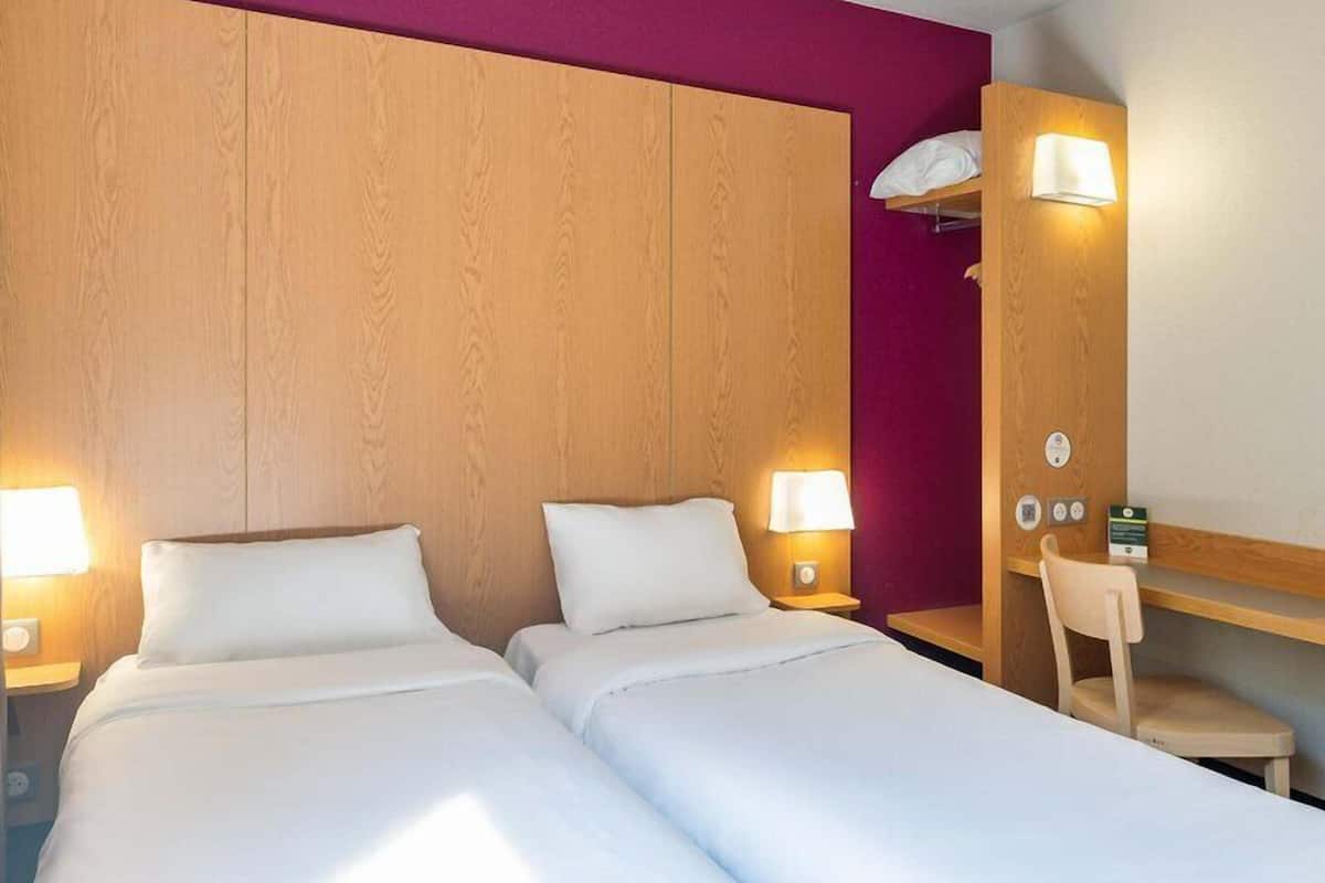 A hotel room with two beds, a desk, a chair, and a wooden headboard.