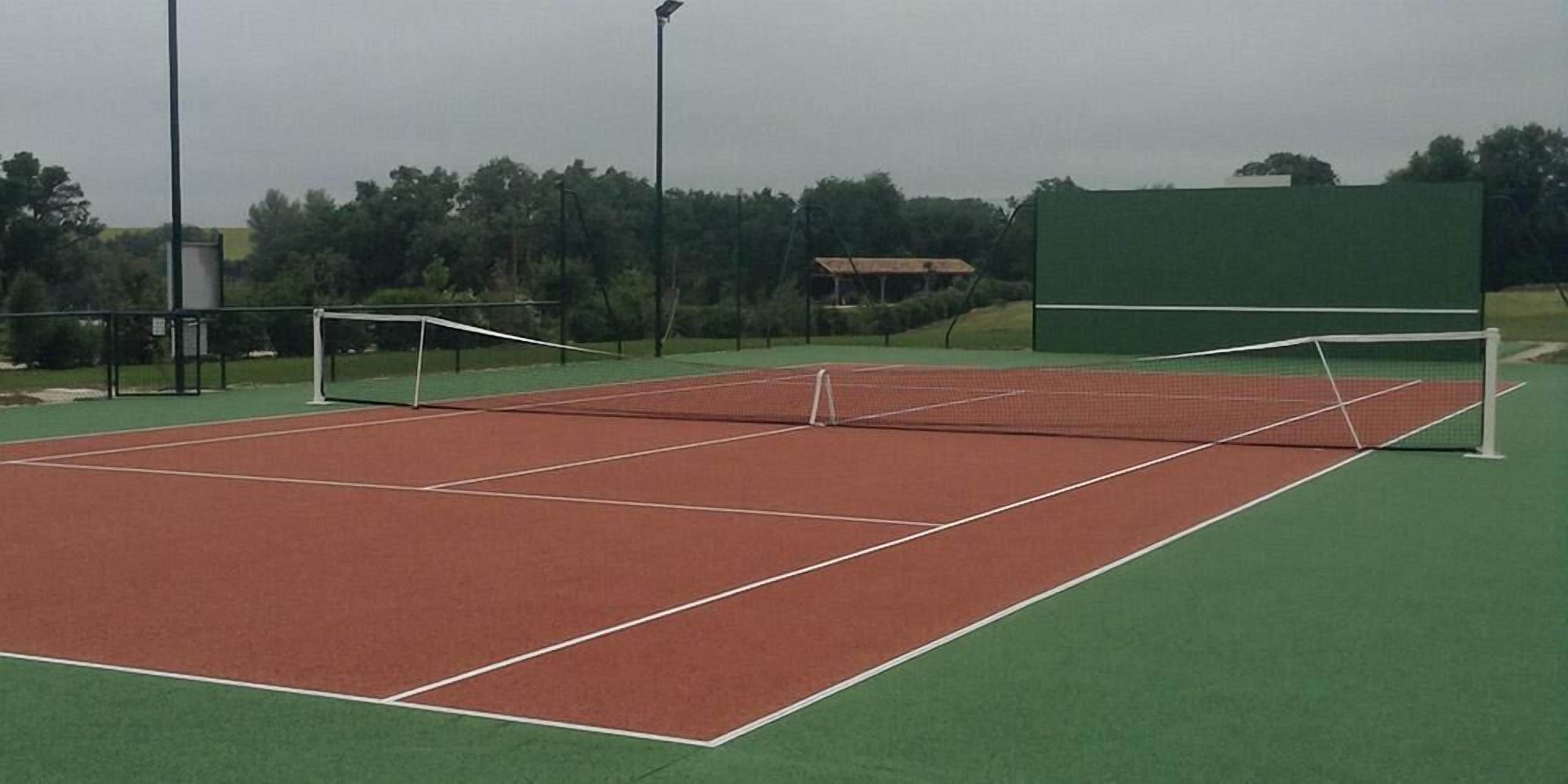 A tennis court with a net, surrounded by green areas and trees.
