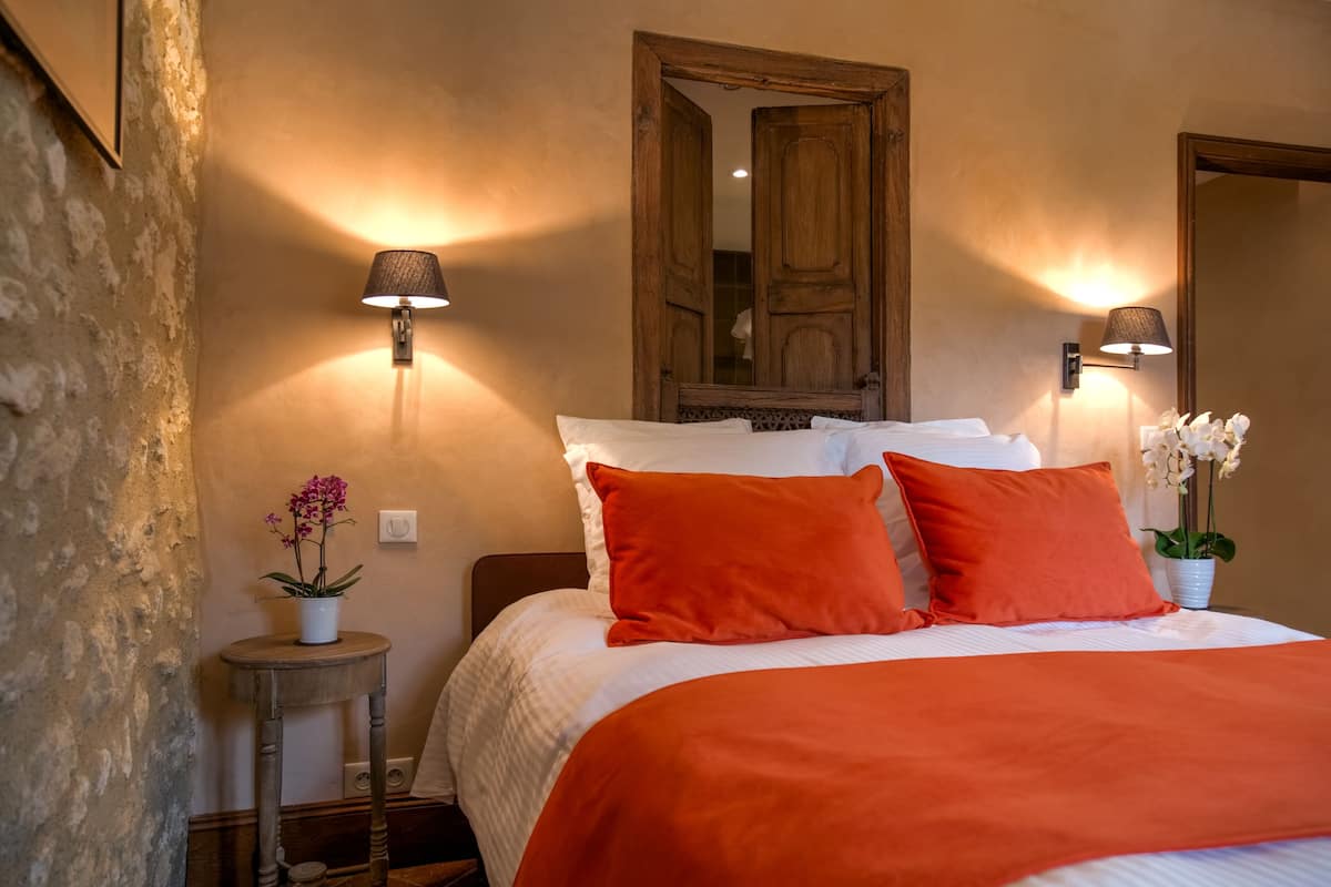 Une chambre à coucher avec un lit, deux lampes fixées au mur, un miroir et une petite table sur laquelle est posée une plante en pot.