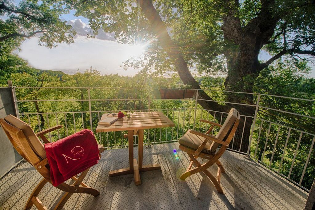 A wooden deck with a table and two chairs, a red towel on one chair, and a view of a forested area.
