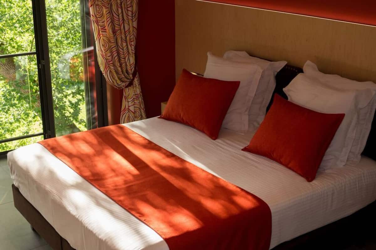 Un lit avec une literie blanche et des oreillers rouges, une fenêtre avec des rideaux et une vue sur les arbres à l’extérieur.