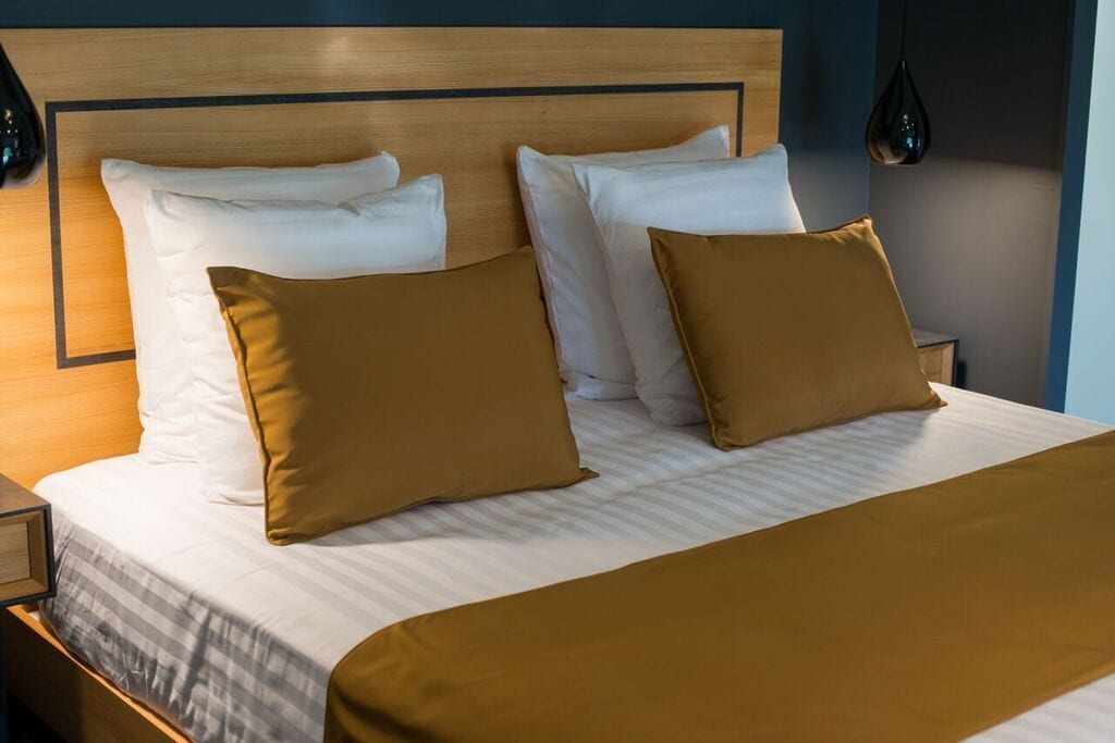 A neatly made bed with white and mustard-colored pillows, a wooden headboard, and a bedside table with a lamp.