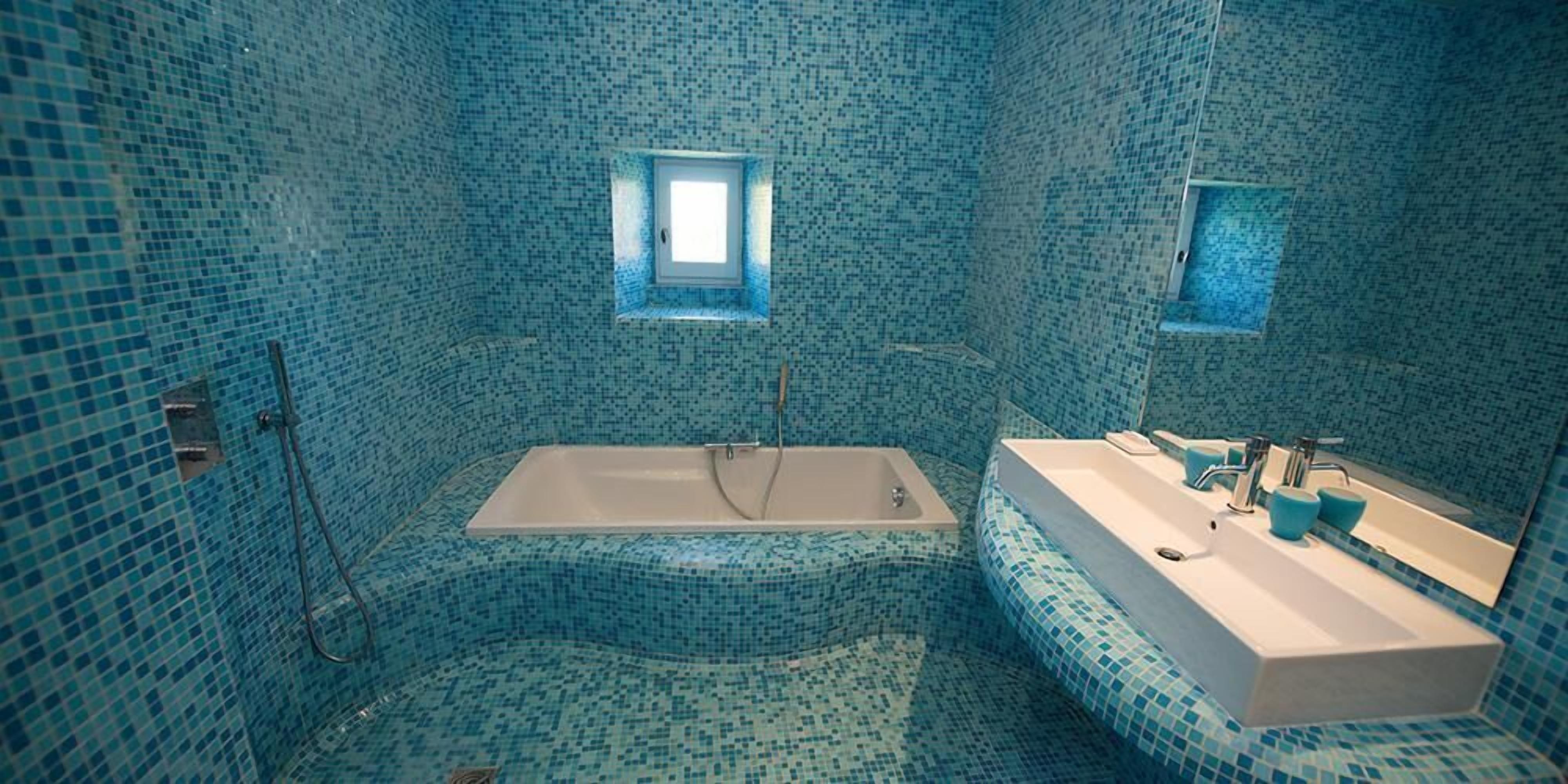 Une salle de bain avec des carreaux de mosaïque bleus, une baignoire, un lavabo avec un miroir et deux fenêtres.