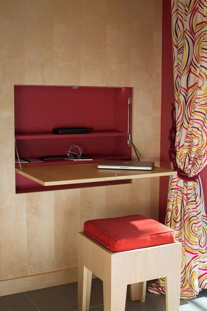 A compact workspace with a laptop on a wooden desk, a red cushioned stool, and a red shelf with a laptop and a small object.