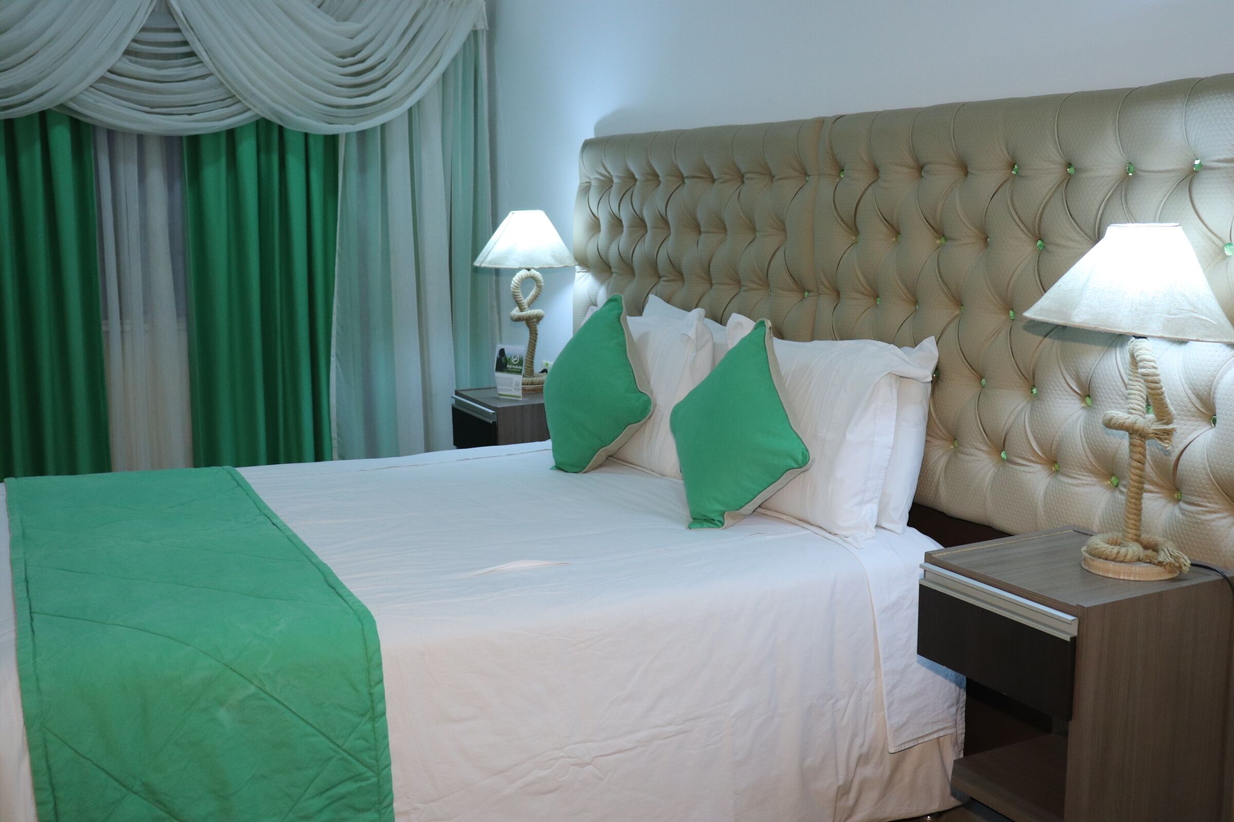 Habitación de hotel con una cama grande, almohadas verdes, cabecero capitonado, dos mesitas de noche con lámparas y una cortina verde.