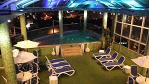 A rooftop pool area with lounge chairs and tables, surrounded by large windows and a glass ceiling. - Hotel Luxsur (Encarnacion)