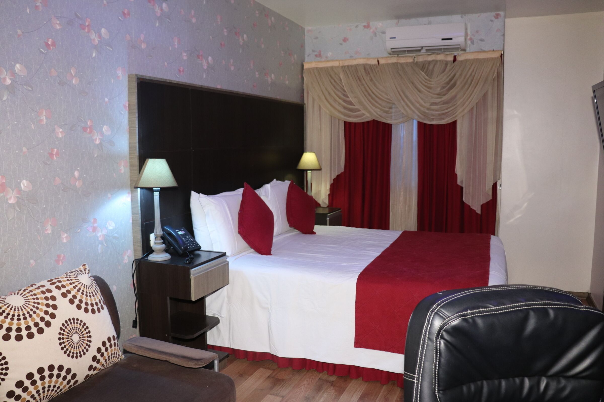A hotel room with a bed, a nightstand with a lamp, a chair, and a red curtain.