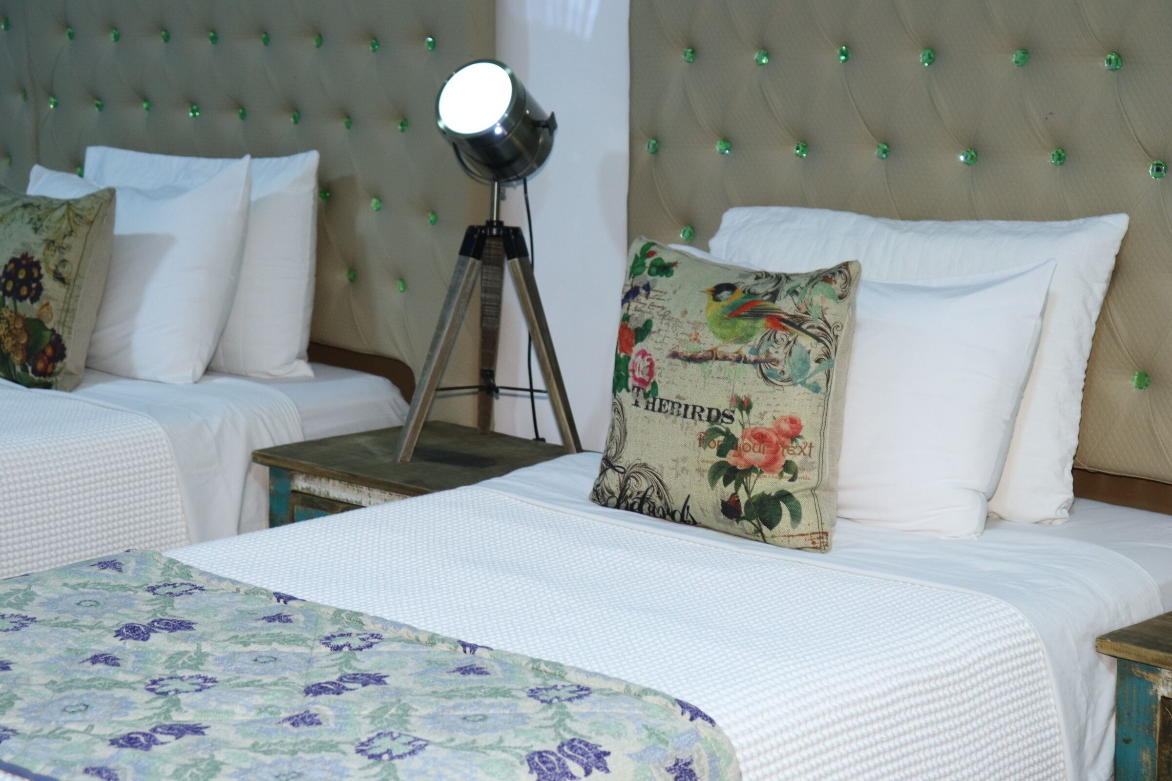 Une chambre d’hôtel avec deux lits : un grand lit recouvert d’une couvre-lit à motifs floraux et un lit plus petit recouvert d’une couvre-lit blanche. On y trouve également une lampe sur pied et un coussin décoratif orné d’un motif d’oiseau.