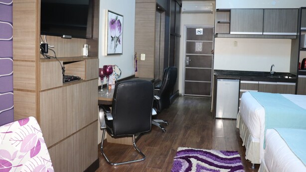 A compact hotel room with a bed, a desk with a chair, a television, a small kitchenette, and a bathroom. - Hotel Luxsur (Encarnacion)