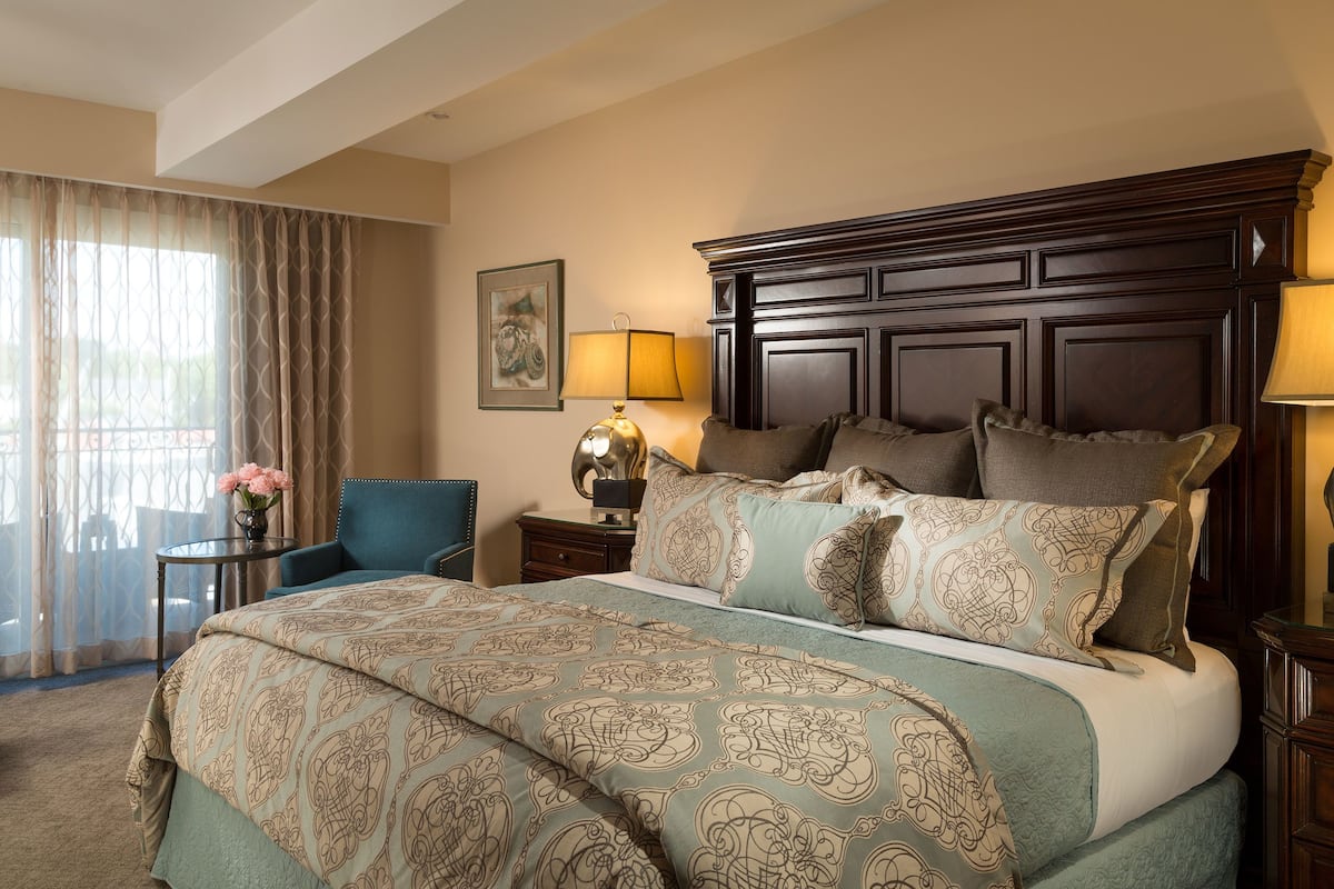 A hotel room with a large bed, a bedside table with a lamp, a chair, and a view of the outdoors through sheer curtains.