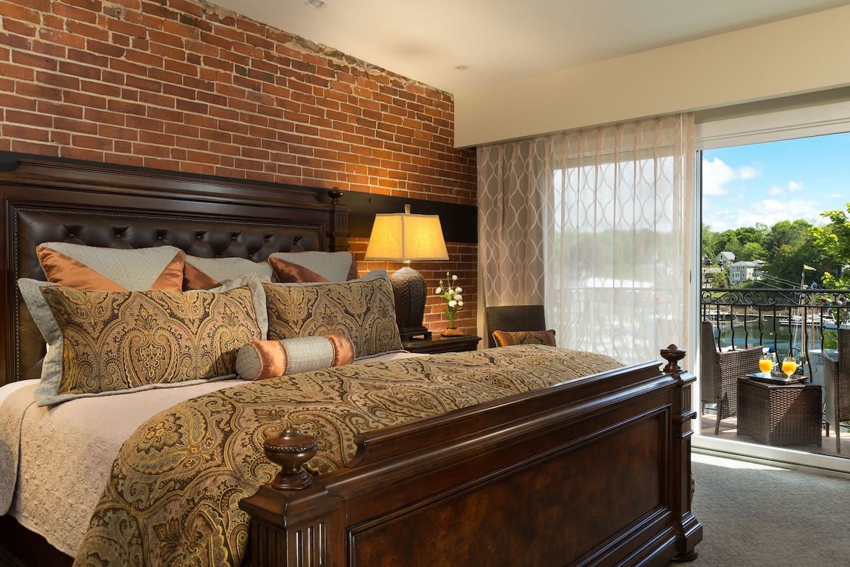 A bedroom with a brick wall, a large bed with patterned bedding, a nightstand with a lamp, and a view of a balcony with outdoor furniture.