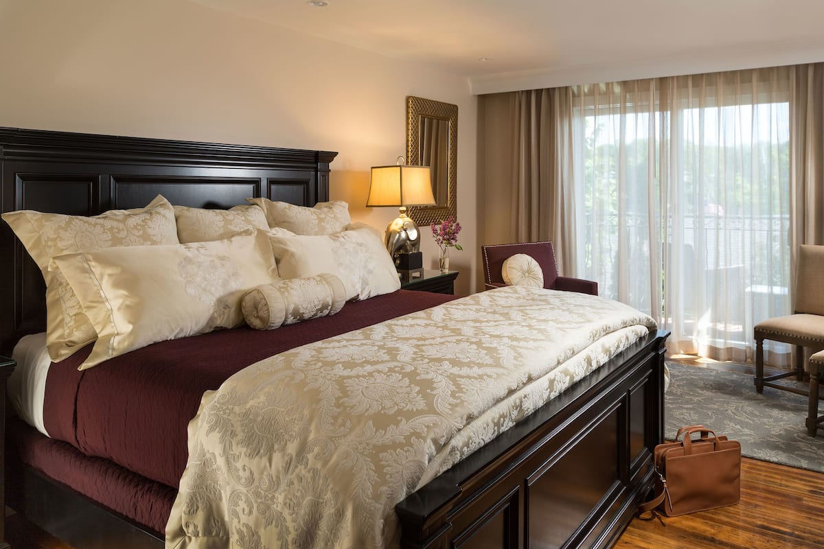 A large bed with a dark headboard, a bedside table with a lamp, a chair, and a view of the outdoors through sheer curtains.