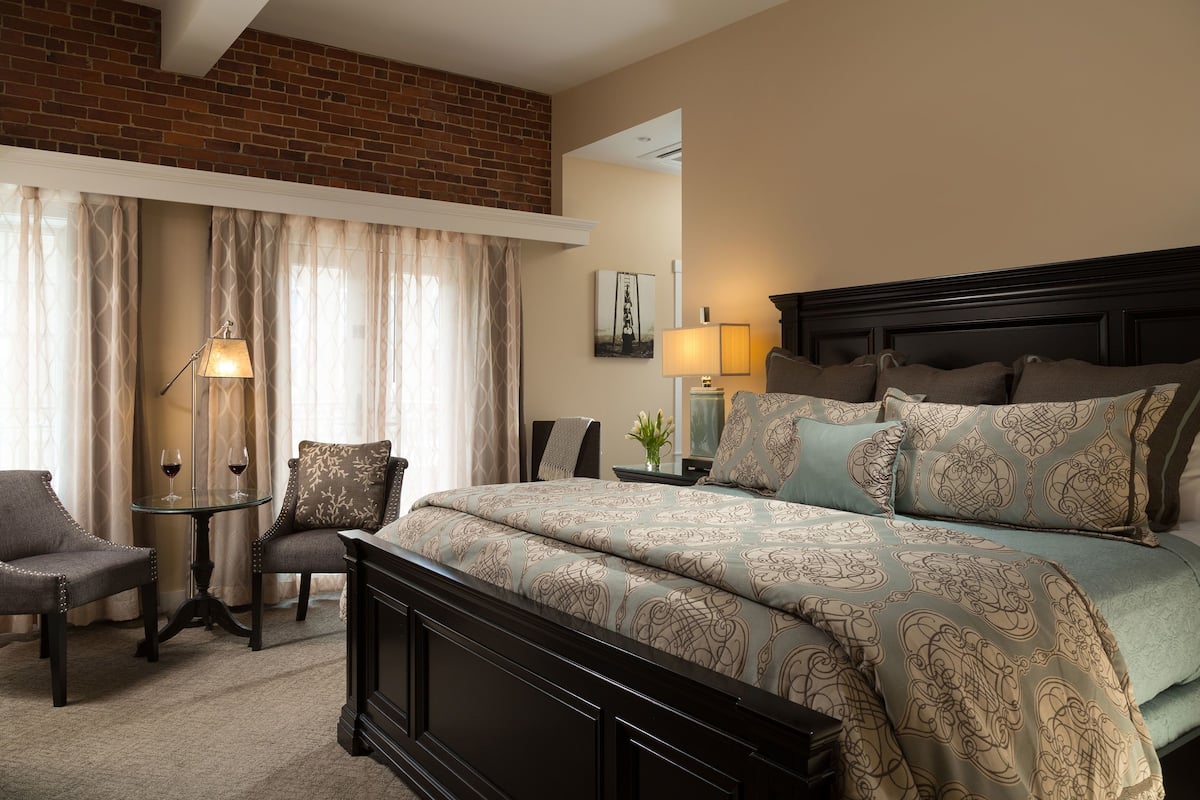 A bedroom with a large bed, two chairs, a small table with a lamp, and a brick wall.