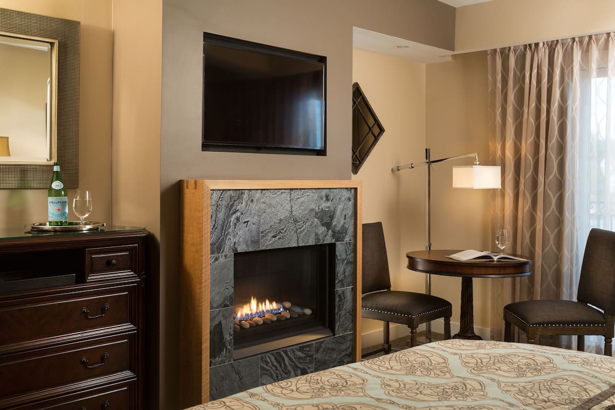 A hotel room with a fireplace, a TV mounted on the wall, a desk with a chair, and a bed with a patterned bedspread.