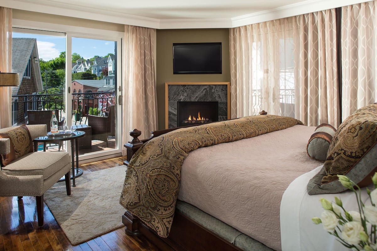 A bedroom with a large bed, a fireplace, a television, and a balcony with a view of houses.