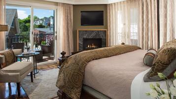 A bedroom with a large bed, a fireplace, a television, and a balcony with a view of houses.