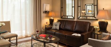 A living room with a leather sofa, a glass coffee table, and a beige rug. There are two lamps on side tables, a vase with flowers, and a framed picture on the wall.