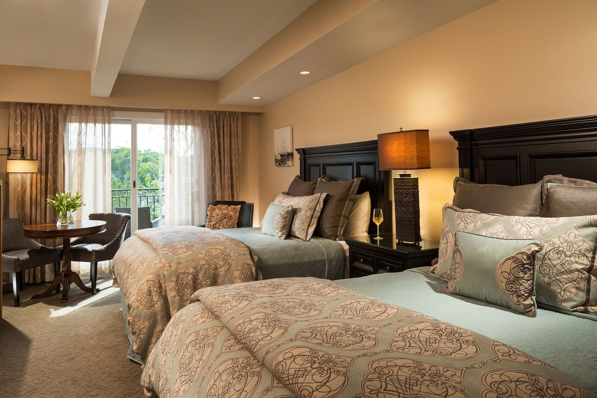 A hotel room with two beds, a sitting area with a table and chairs, and a view of the outdoors through large windows.
