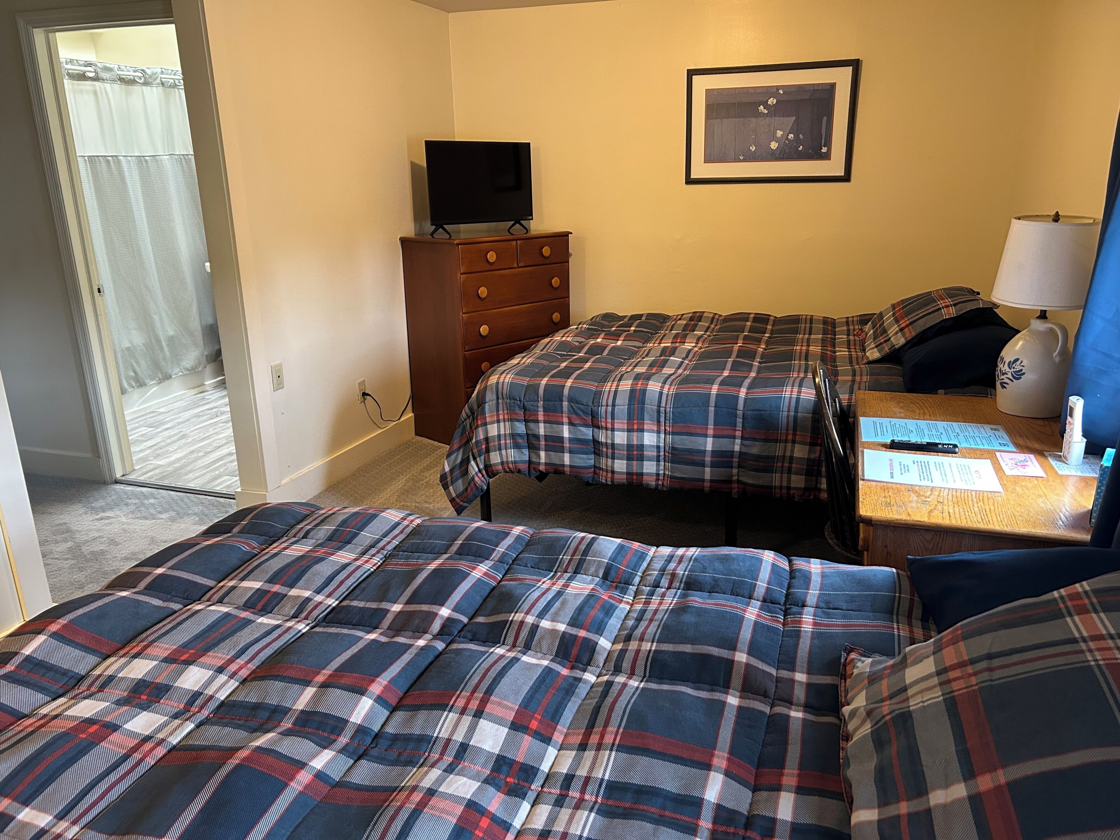 A hotel room with two beds, a TV on a dresser, a framed picture on the wall, and a desk with papers and a lamp.