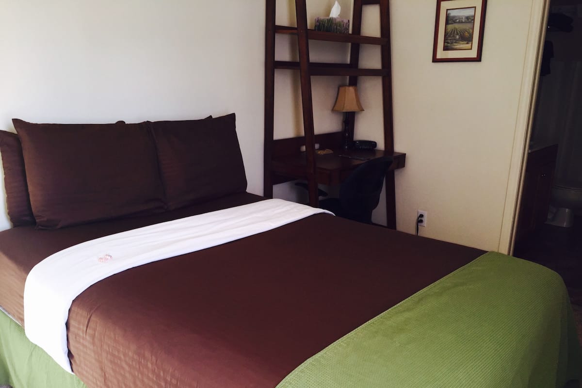 A neatly made bed with brown and green bedding, a desk with a lamp, and a framed picture on the wall.