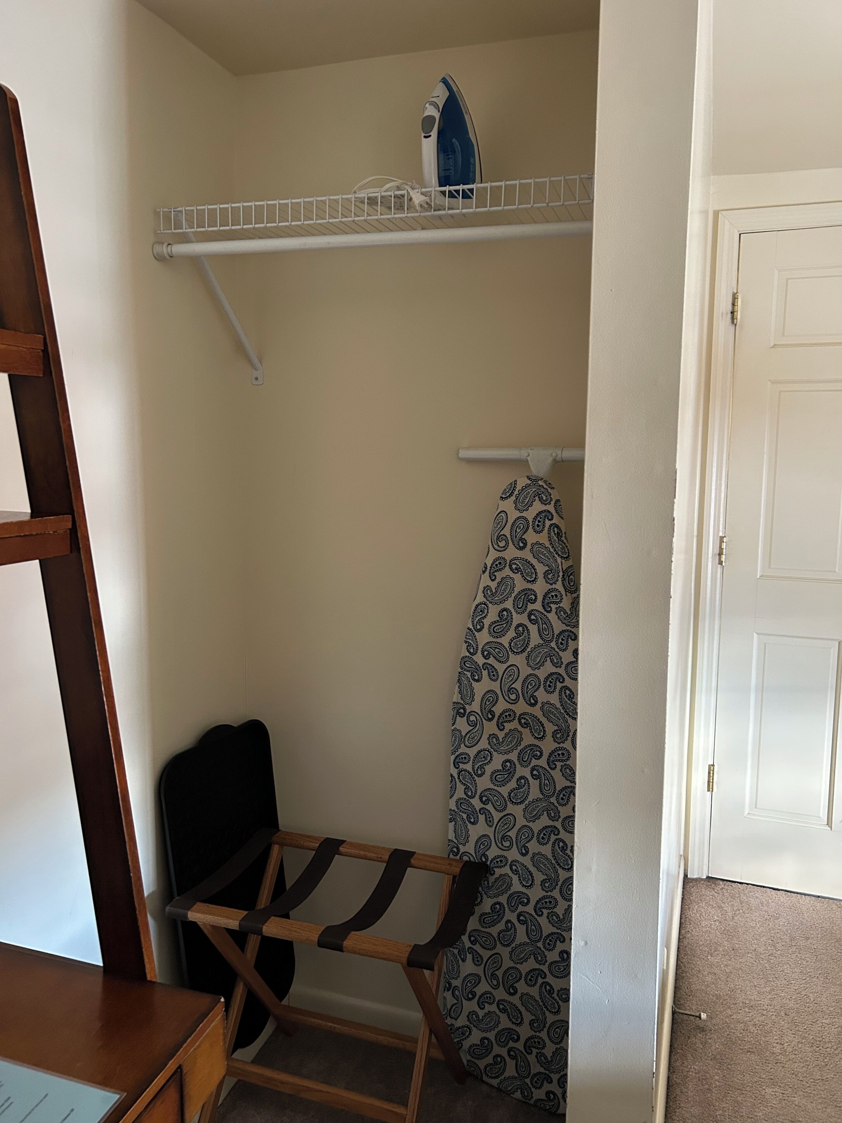 A room with a wooden chair, a shelf with an iron, and a patterned item hanging on the wall.