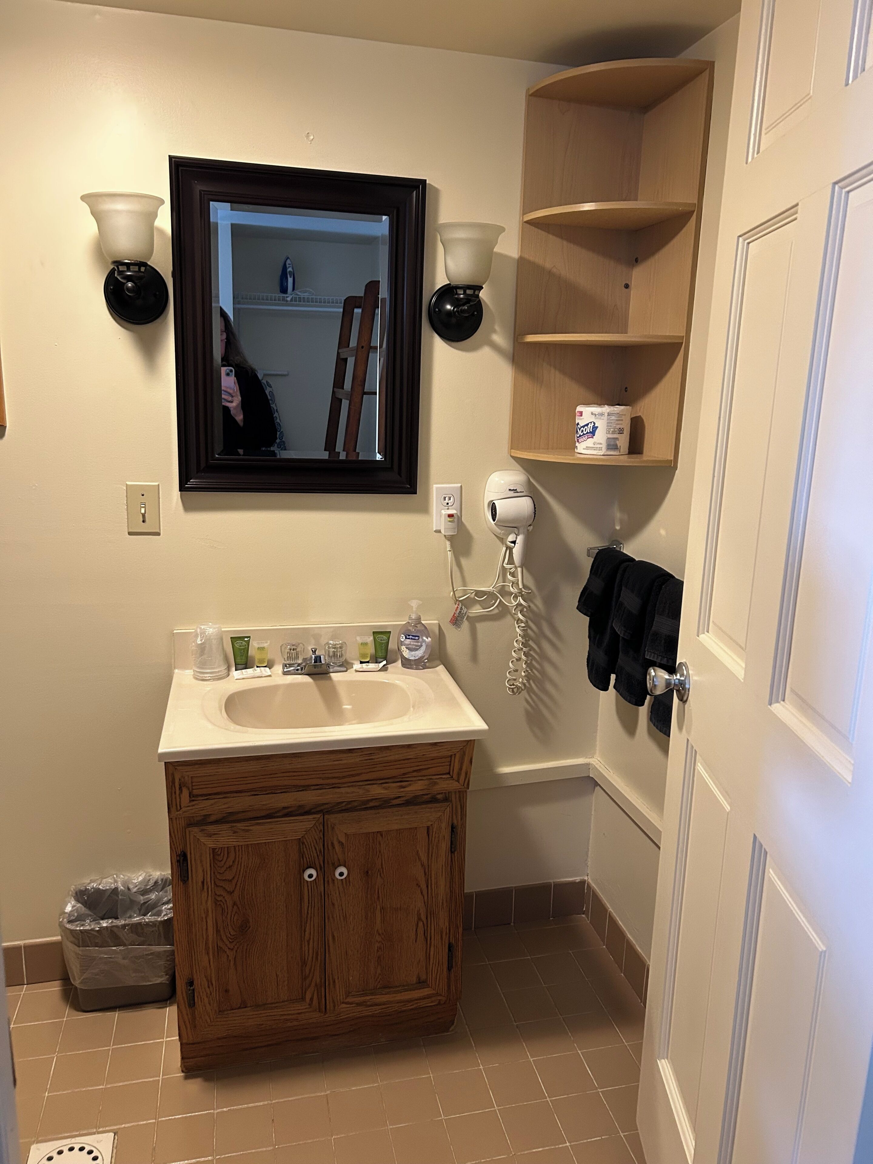 Un baño con tocador de madera, un espejo, dos luces empotradas, un perchero para toallas y un estante con artículos de aseo.