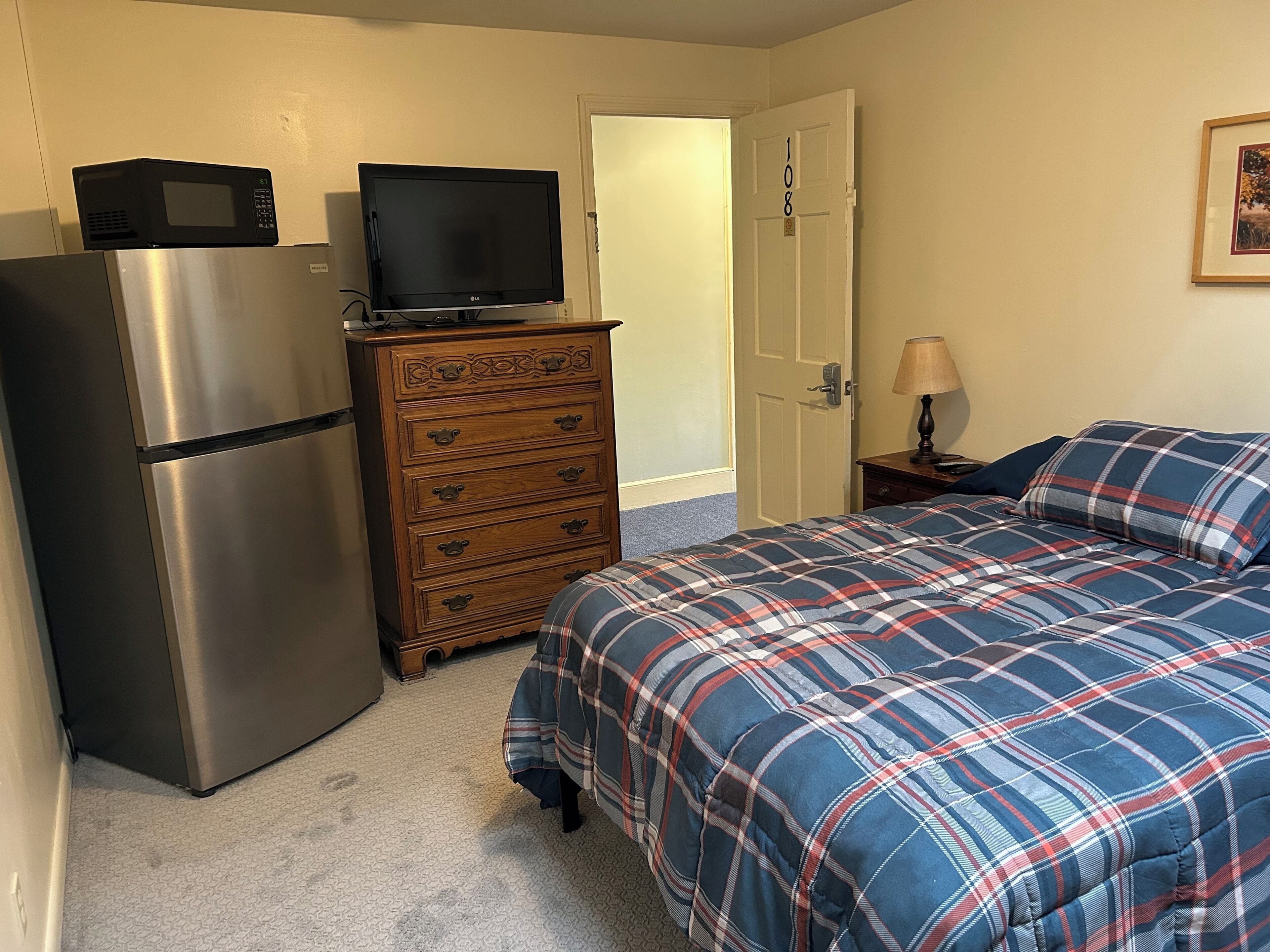 Une chambre à coucher avec un lit, une commode, un téléviseur posé sur un réfrigérateur et une porte donnant sur une autre pièce.