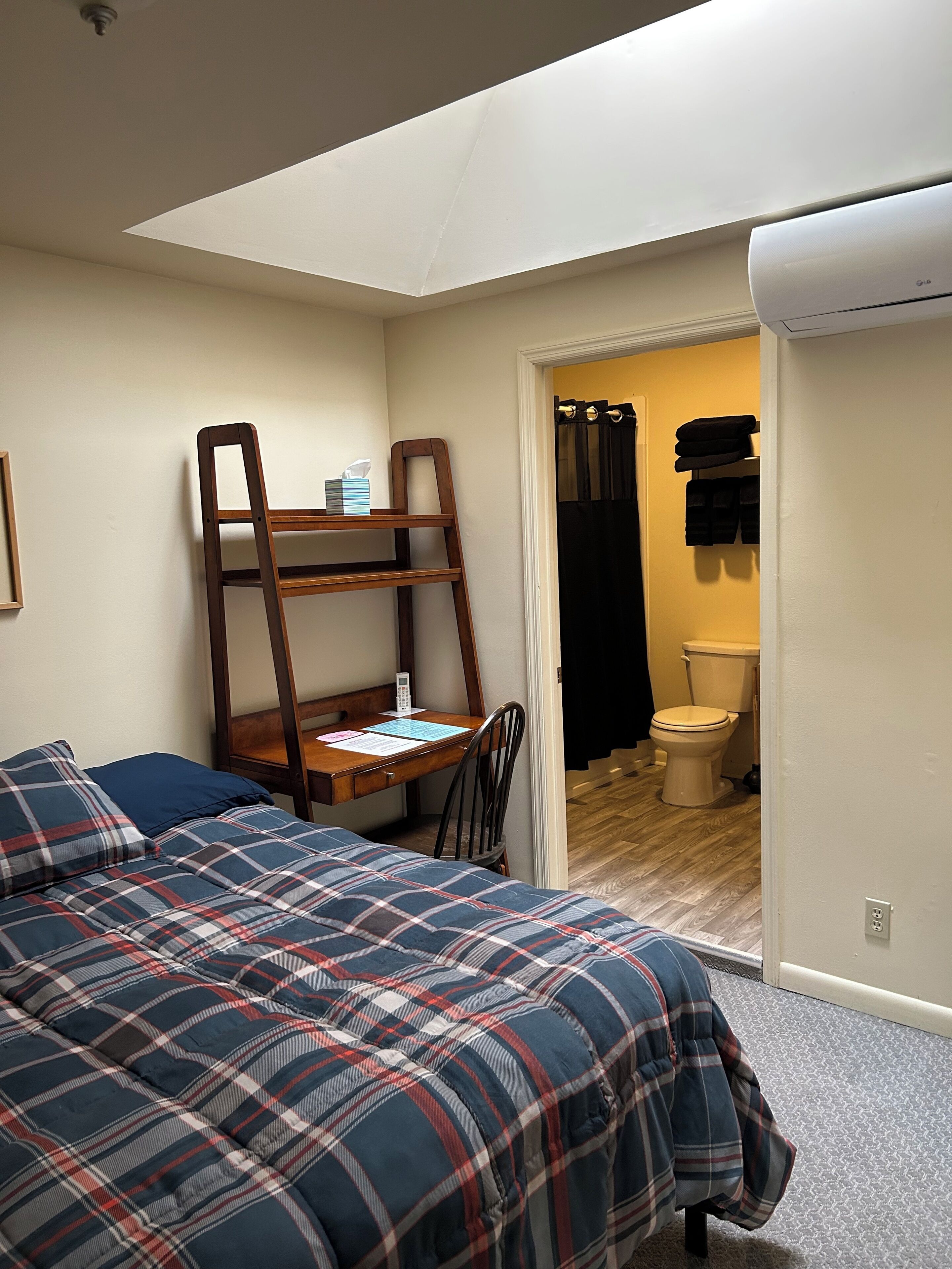 Quarto com cama, escrivaninha e cadeira. Há uma janela sobre a escrivaninha, e um banheiro visível através de uma porta aberta.