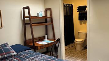 A bedroom with a bed, a desk, and a chair. There is a window above the desk, and a bathroom is visible through an open door.