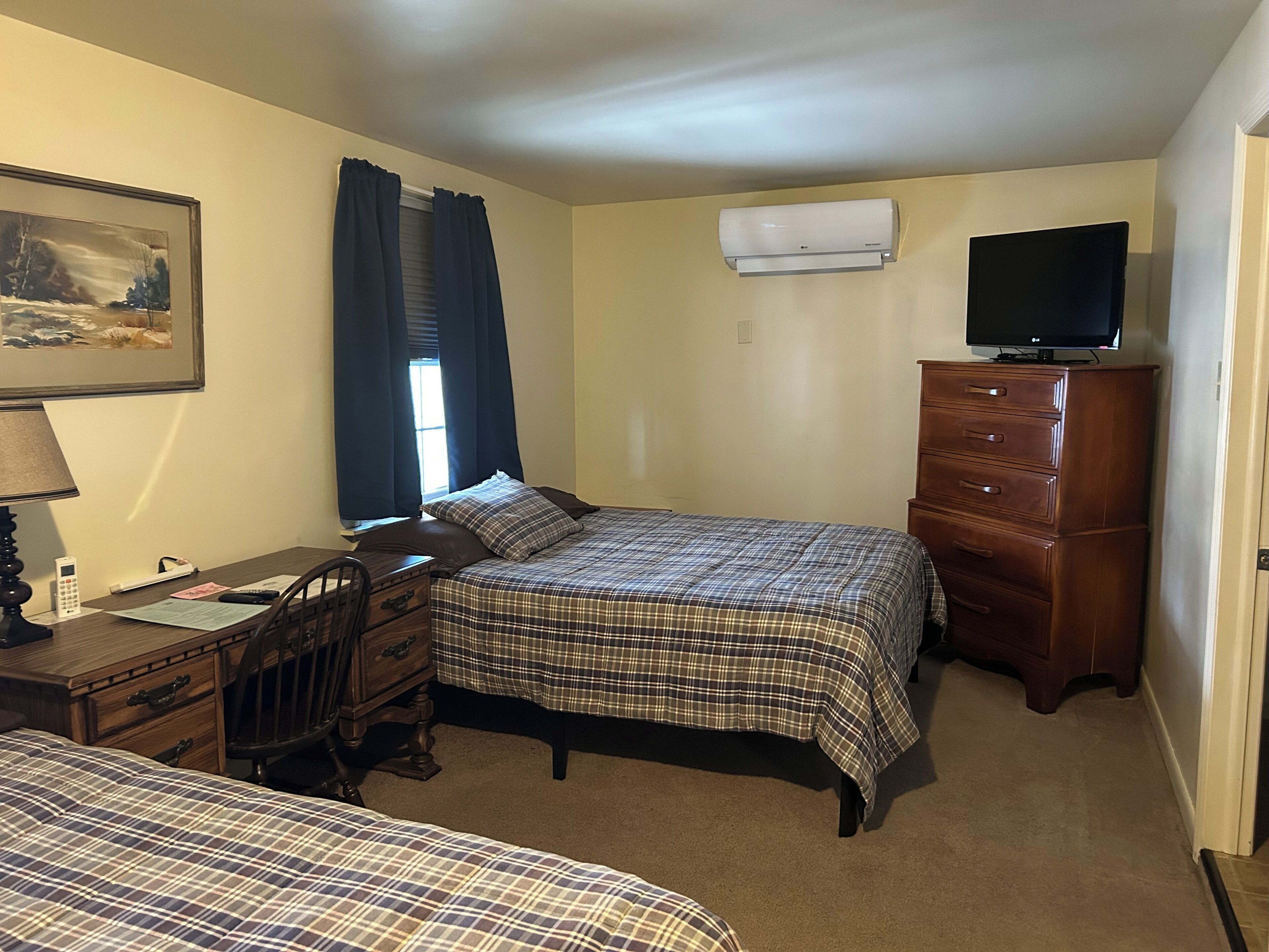 A bedroom with two beds, a dresser, a desk with a lamp, a television, and an air conditioner.