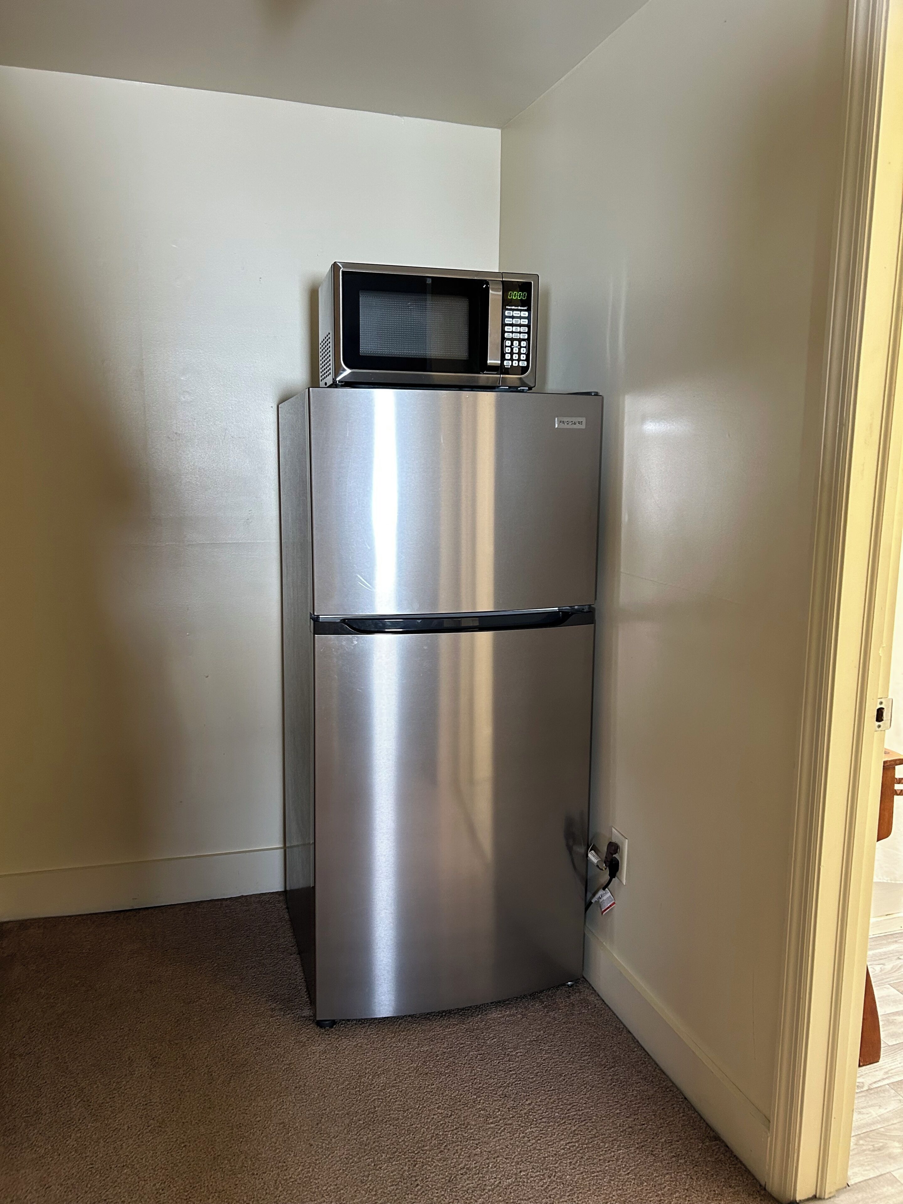 Een roestvrijstalen koelkast met een magnetron erboven, in een hoek van een kamer met beige wanden en vloerbedekking.