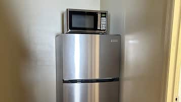 Een roestvrijstalen koelkast met een magnetron erboven, in een hoek van een kamer met beige wanden en vloerbedekking.