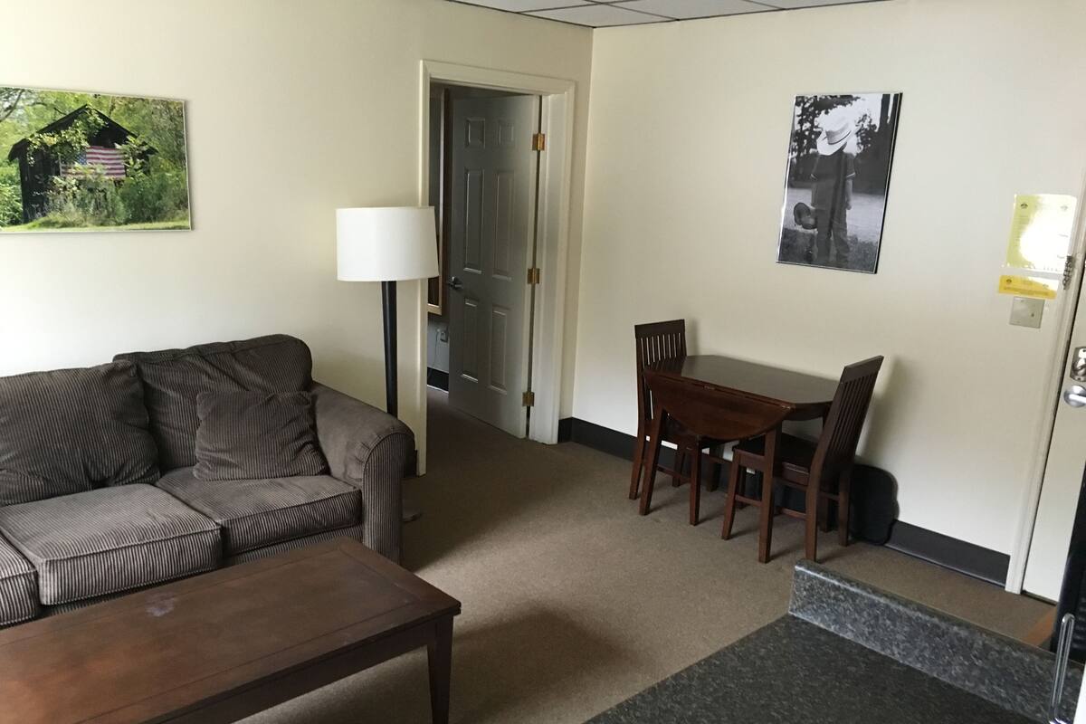 A living room with a sofa, a coffee table, a dining table with chairs, a lamp, and a door leading to another room.