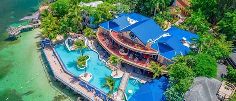 An aerial view of a resort with a large pool, multiple decks, and lush greenery.
