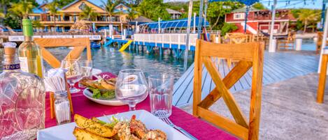 A dining table with a plate of seafood, glasses, and a bottle of wine, set on a wooden deck with a view of a water body and buildings in the background.