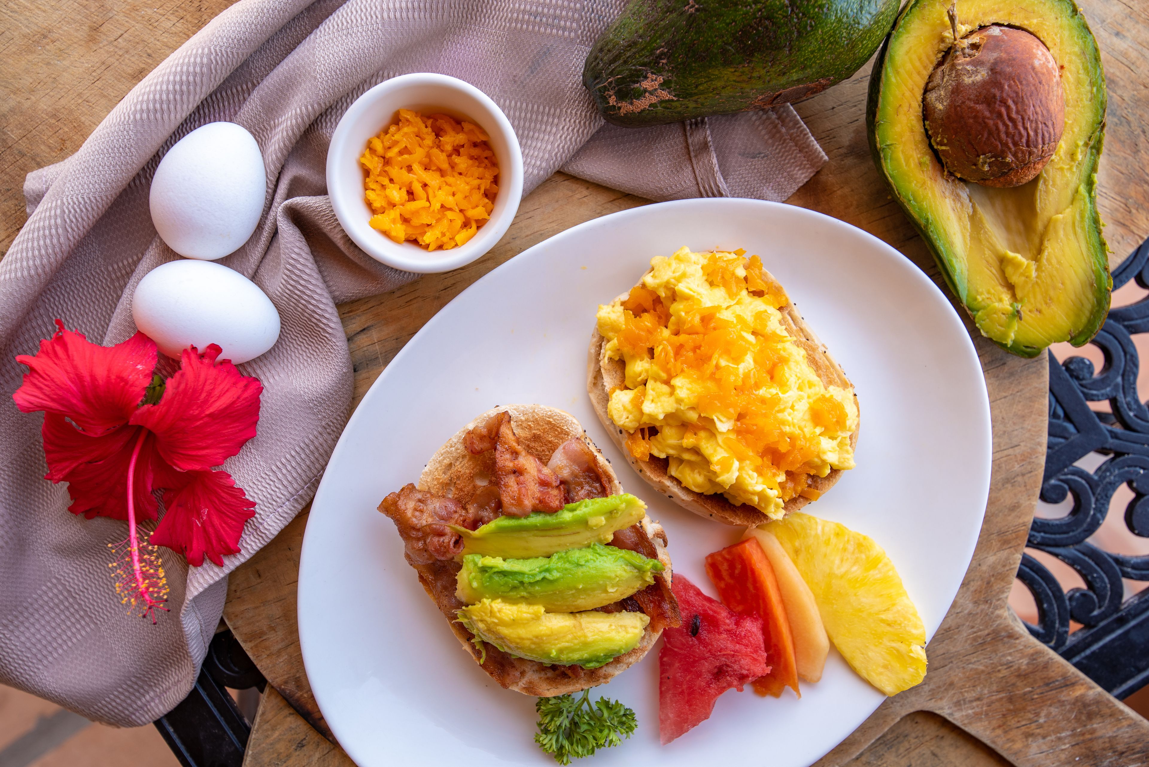 A breakfast plate with eggs, avocado, bacon, and fruit on a wooden table.