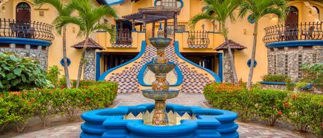 A building with a fountain in the center, surrounded by palm trees and red flowers.