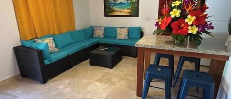 A living room with a sectional sofa, a ceiling fan, a painting of a beach, and a kitchen area with stools and a flower vase.