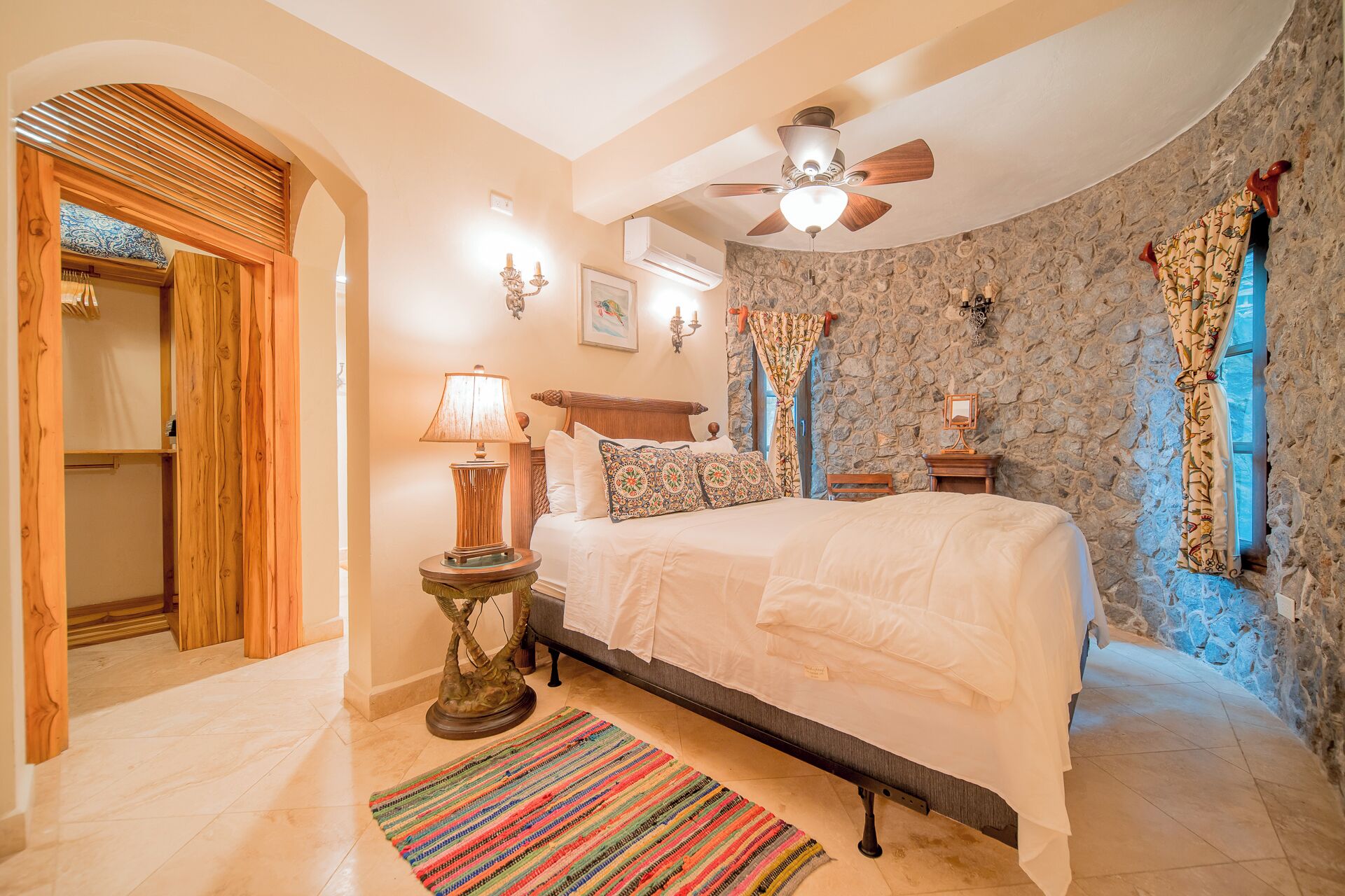 Un dormitorio con cama, ventilador de techo, pared de piedra y puerta de madera.