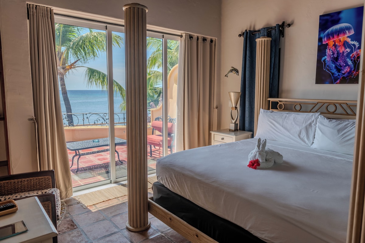 Habitación de hotel con una cama, una mesita de noche, una lámpara y vista al océano a través de amplios ventanales.