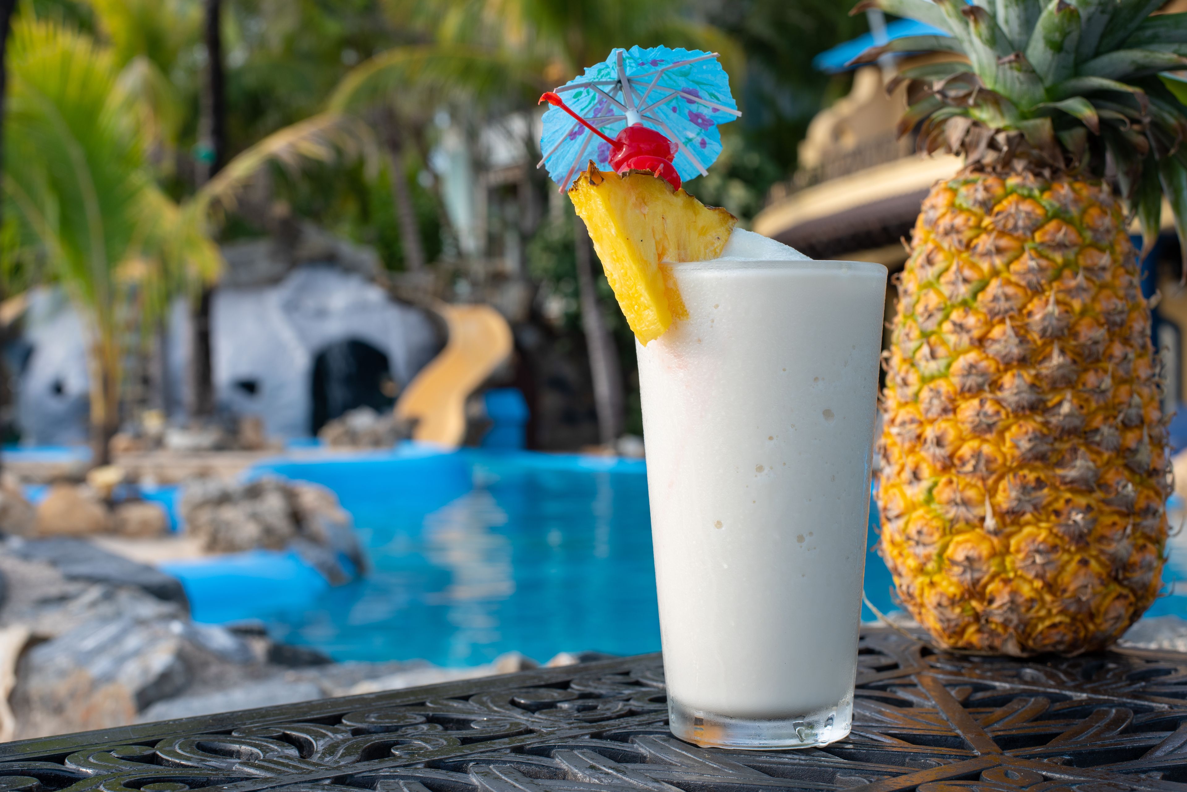 A glass of a creamy beverage with a pineapple slice and a decorative umbrella on the rim, placed on a metal table beside a pineapple, with a pool and palm trees in the background.