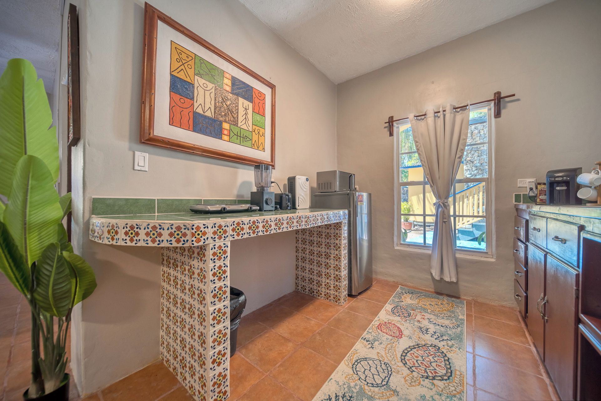 A kitchen with a tiled backsplash, a refrigerator, a countertop with appliances, and a window with curtains.