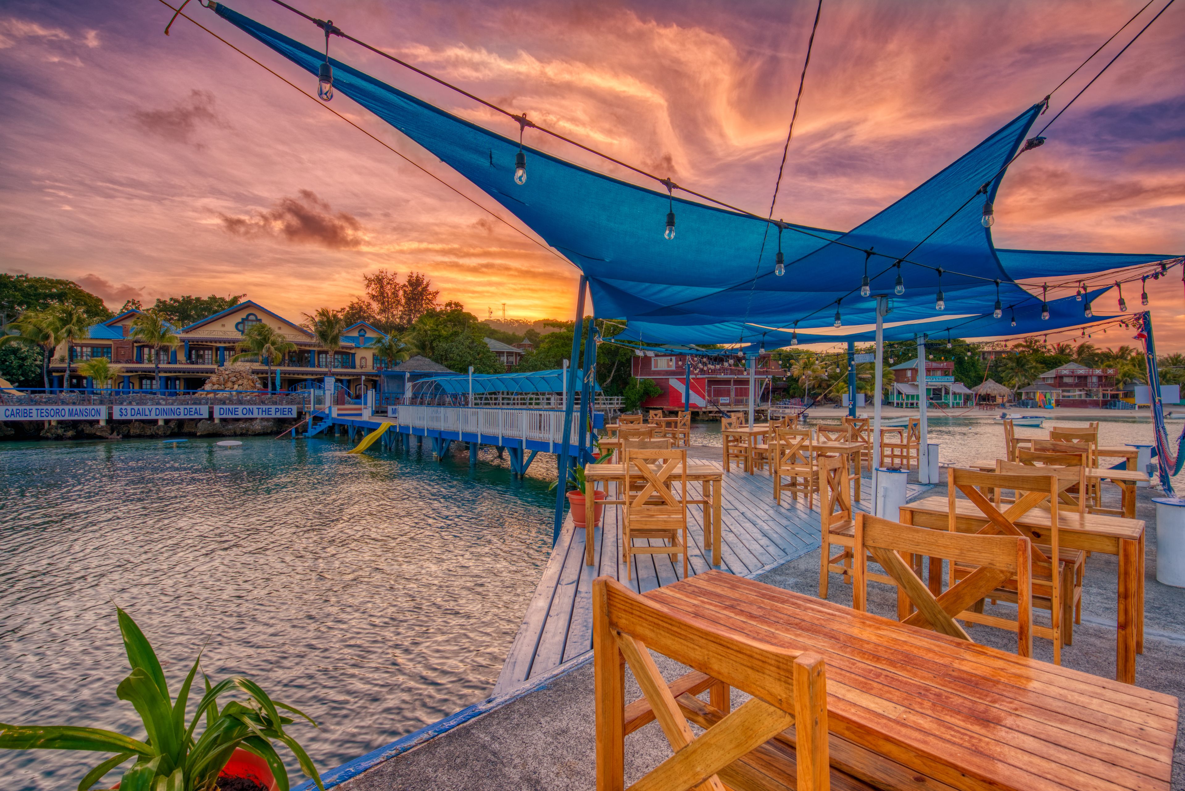 A waterfront restaurant with outdoor seating, a pier, and a sunset view.