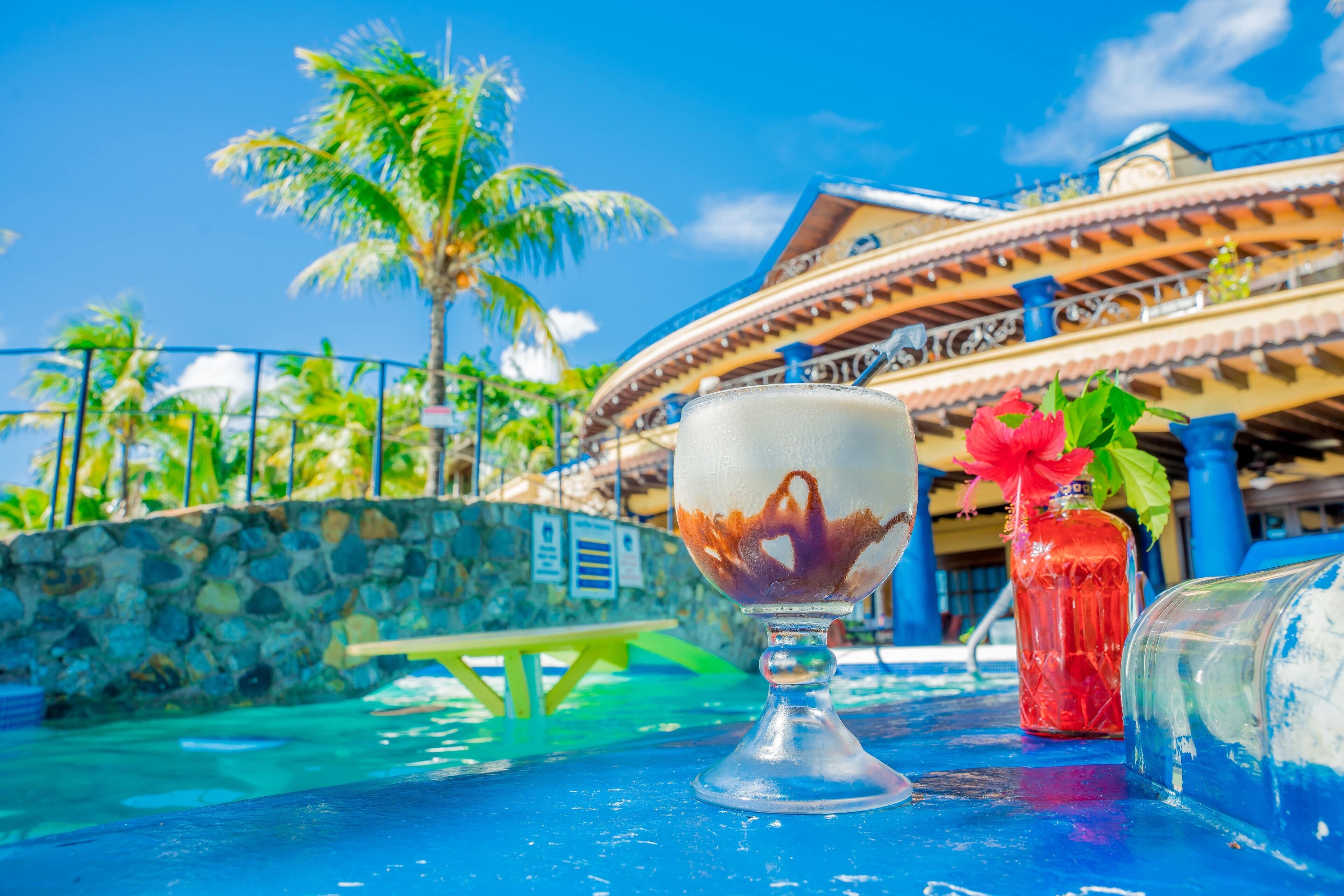 A poolside setting with a clear glass bowl containing a drink, a red bottle with a flower, and a blue glass.