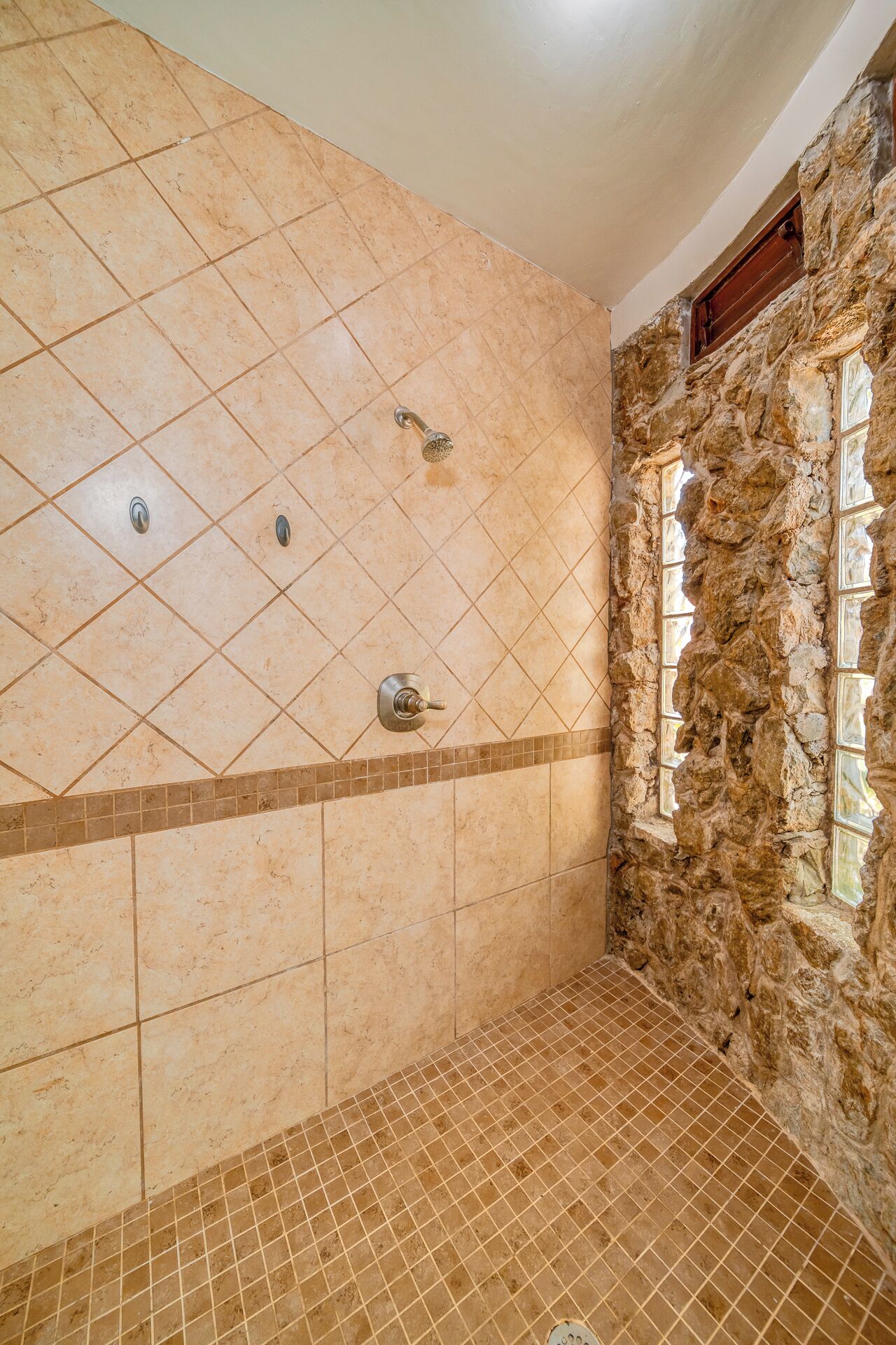 Casa de banho com azulejos bege, chuveiro com parede de pedra e janela.