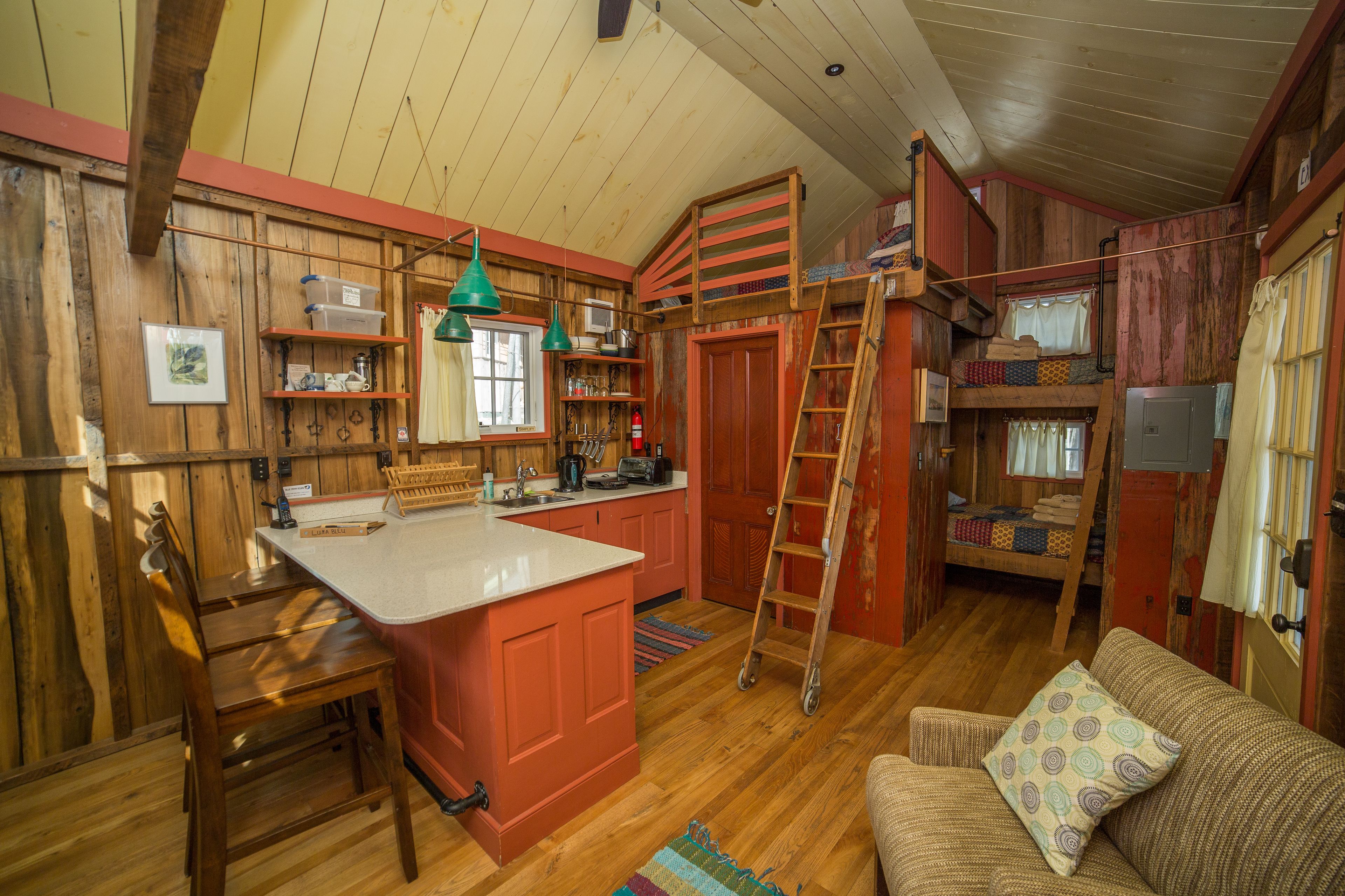 A cozy cabin interior with a wooden interior, a kitchen area with a central island, a bunk bed, and a sofa with a patterned cushion.