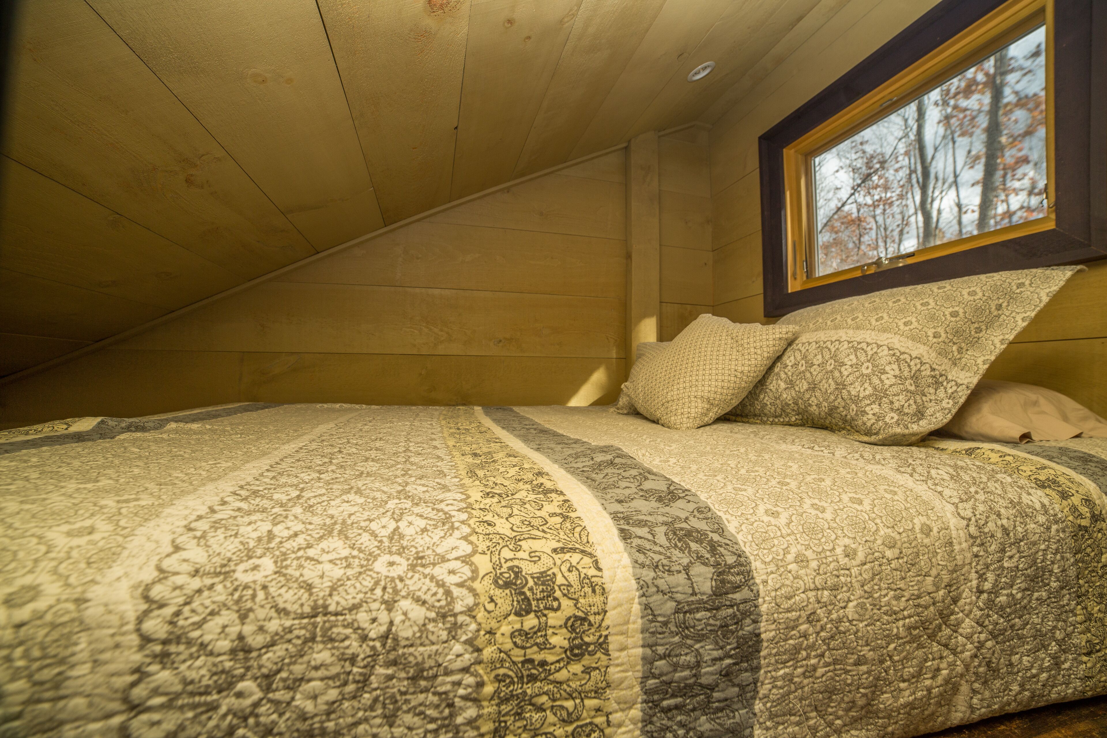 Een knus slaapkamer op zolder met een schuine wand, een groot raam en een bed met beddengoed met een patroon.