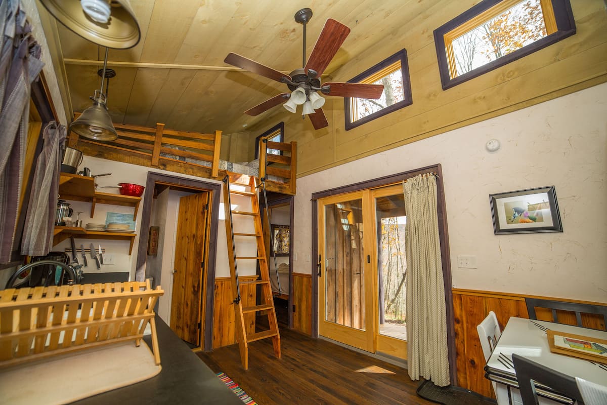 A rustic cabin interior with a loft bed, wooden furniture, a ceiling fan, and a dining area.